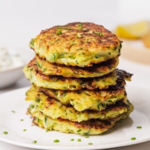 A stack of golden-brown zucchini fritters garnished with chopped chives sits on a white plate. A bowl of dip and lemon wedges are blurred in the background.