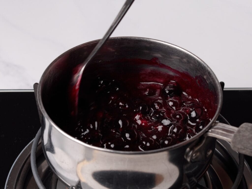 A spoon stirs a dark, glossy blueberry sauce in a stainless steel saucepan on a stovetop.