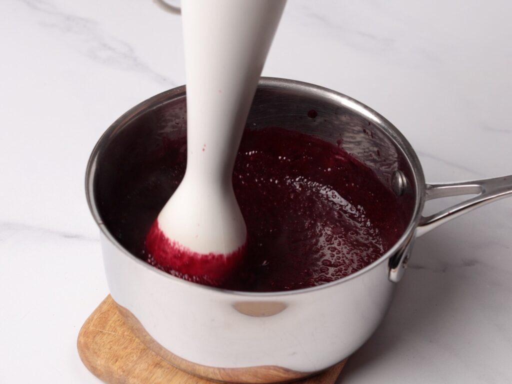 A white immersion blender is blending a dark purple mixture in a stainless steel saucepan, which sits on a wooden trivet against a white background.