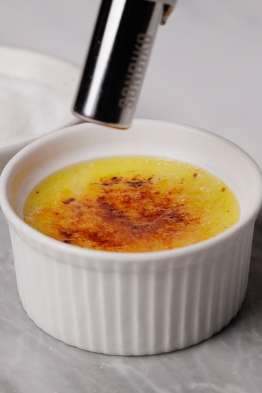 A kitchen torch caramelizes the sugar on top of crème brûlée in a white ramekin, creating a golden, crispy layer.