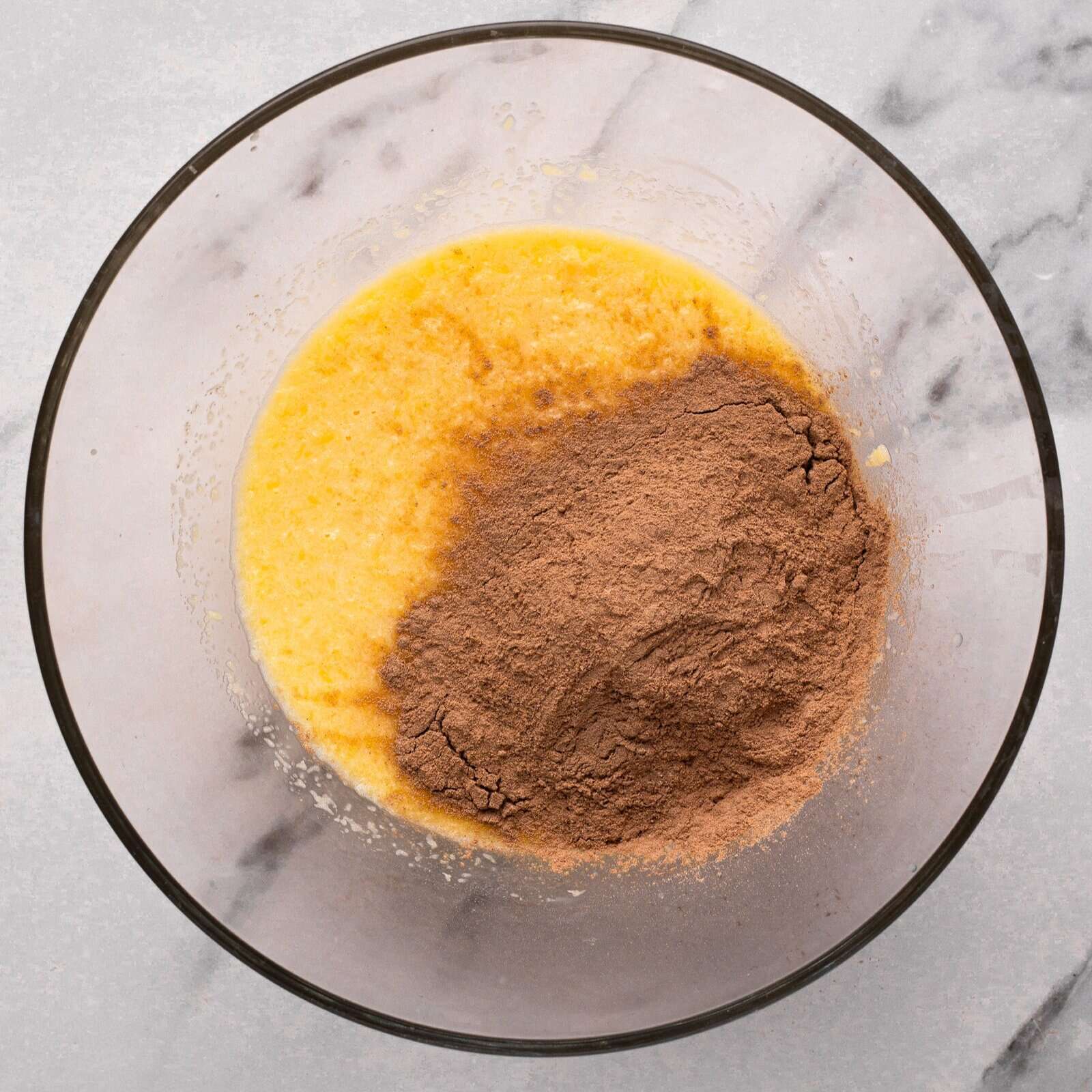 A glass bowl on a marble surface contains a yellow batter with a mound of cocoa powder on top, not yet mixed in.