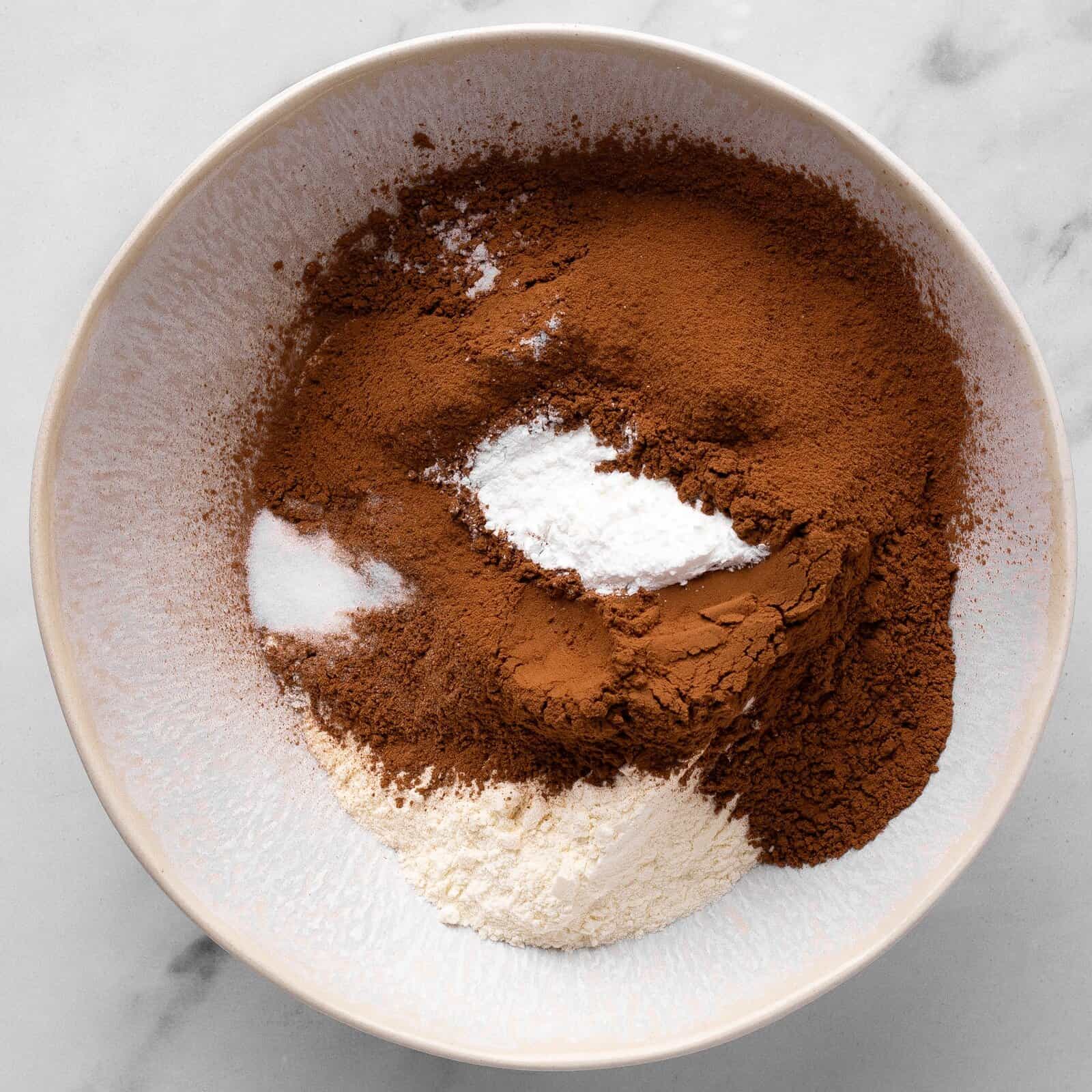 A white ceramic bowl on a marble surface holds piles of cocoa powder, flour, baking powder, and salt, arranged separately before mixing.