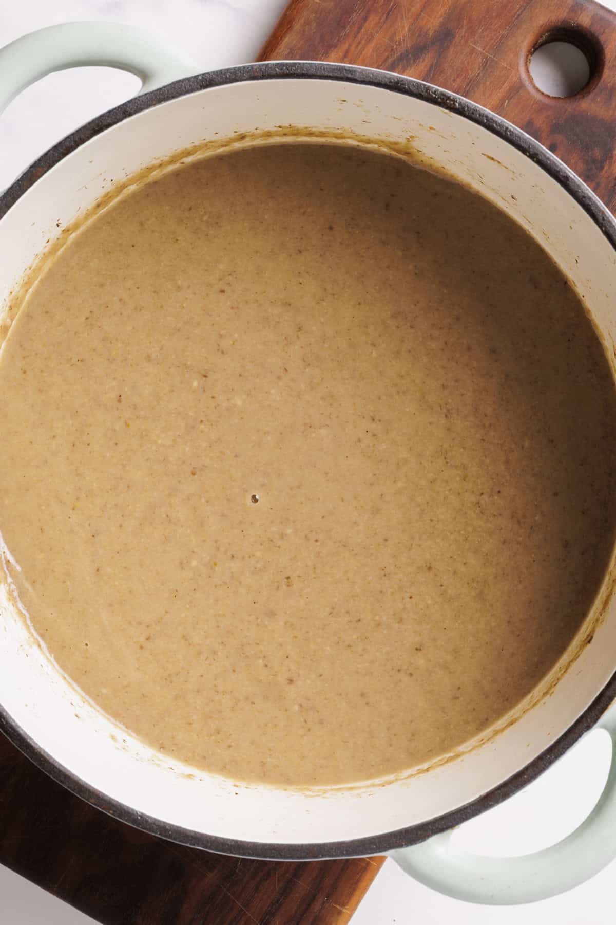 A large white pot filled with creamy brown mushroom soup sits on a wooden cutting board, viewed from above.