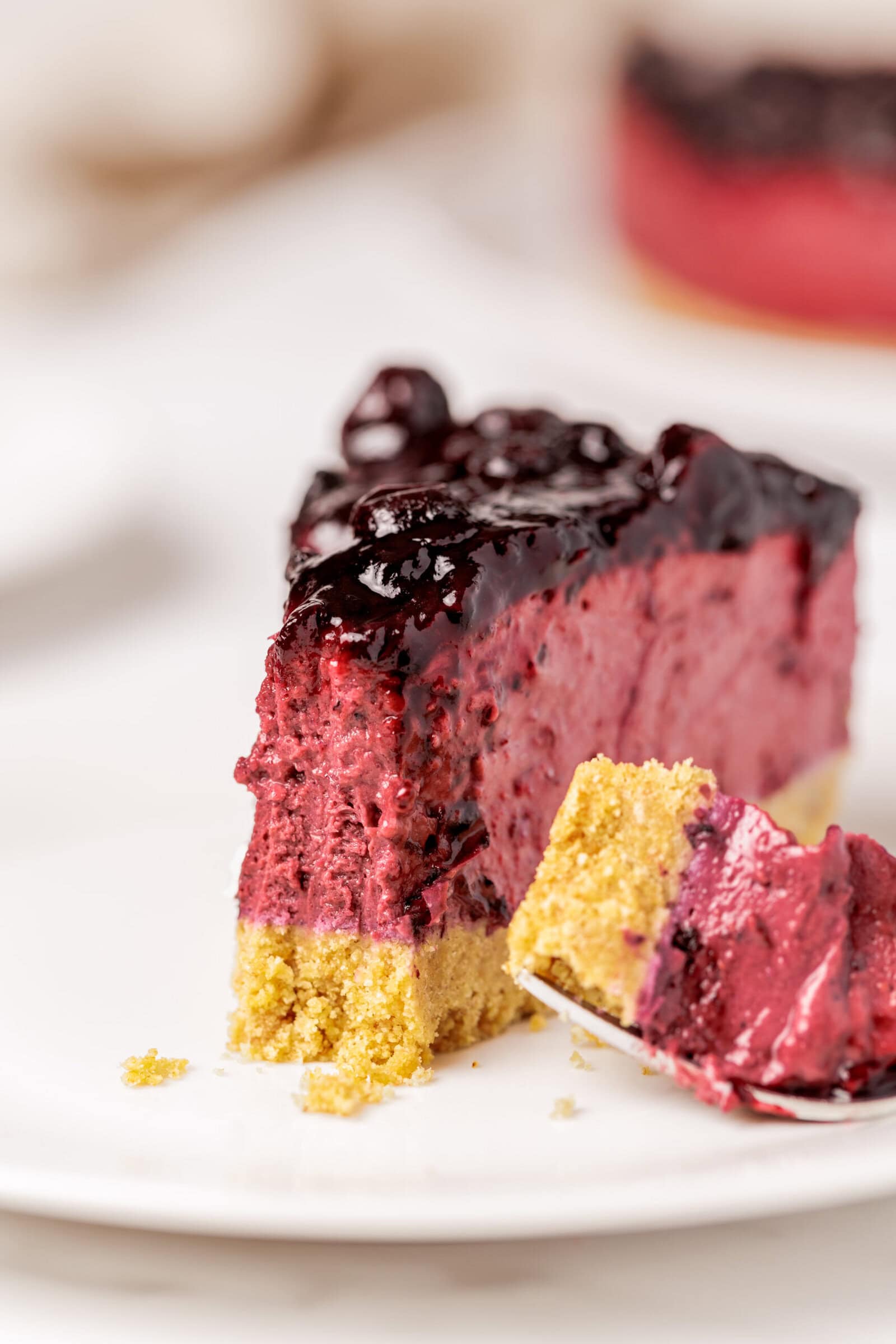 A close-up of a slice of a no-bake blueberry cheesecake with a golden crust, topped with a glossy berry sauce, sitting on a white plate. A fork holds a bite of cheesecake and crust in the foreground.