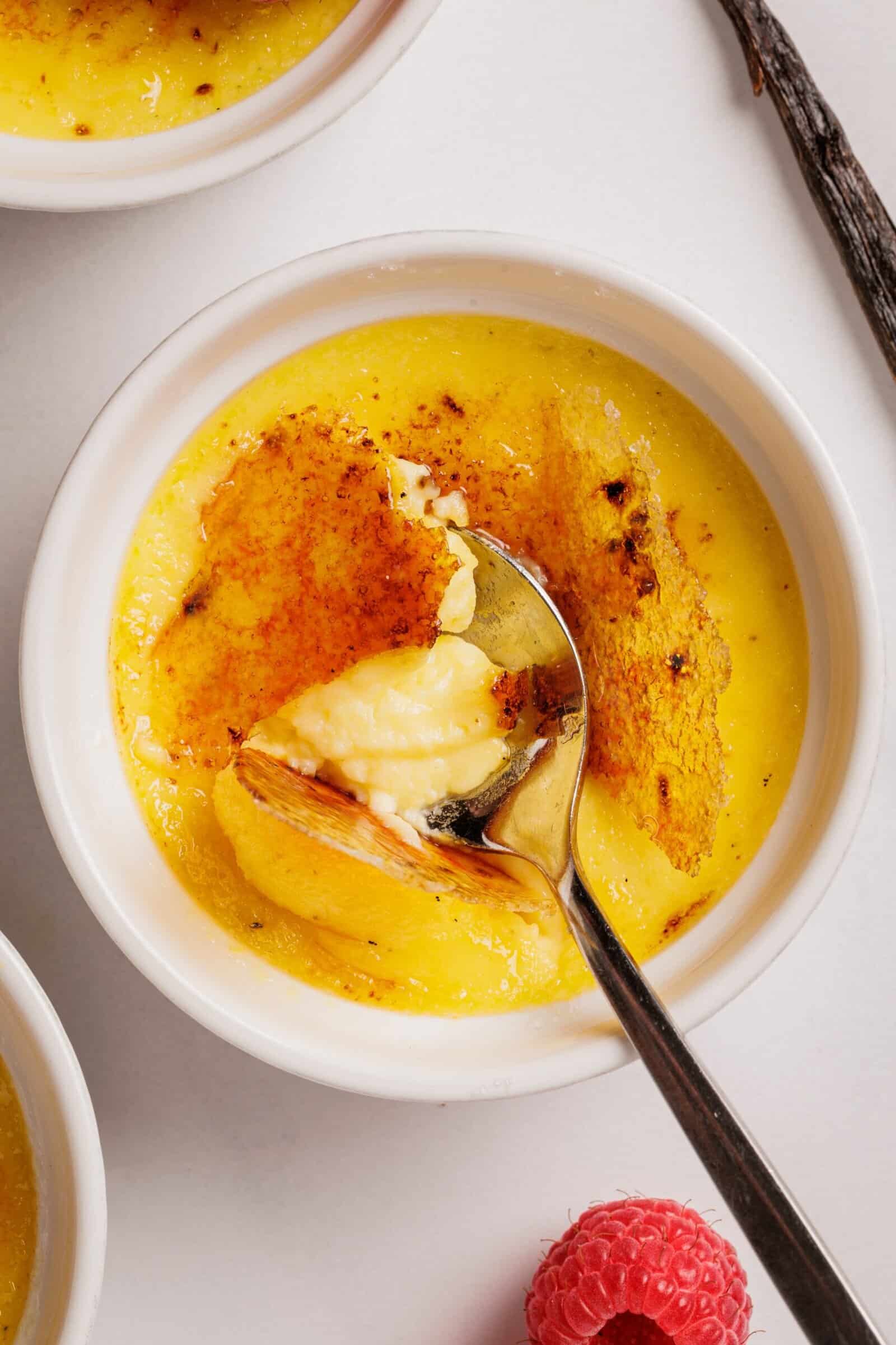 A spoon breaking through the caramelized sugar crust of a crème brûlée in a white ramekin, revealing the creamy custard inside. A raspberry and a vanilla bean are nearby on a white surface.