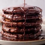 A tall stack of chocolate pancakes on a plate, covered in rich chocolate sauce that is being poured over the top, dripping down the sides.