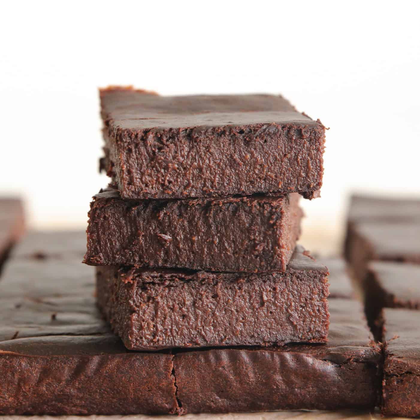Three thick, rich gluten-free healthy chocolate brownies that are made with 3 ingredients are stacked on top of each other, with more brownie pieces visible on the table around them. The texture appears dense and fudgy, and the background is softly blurred.