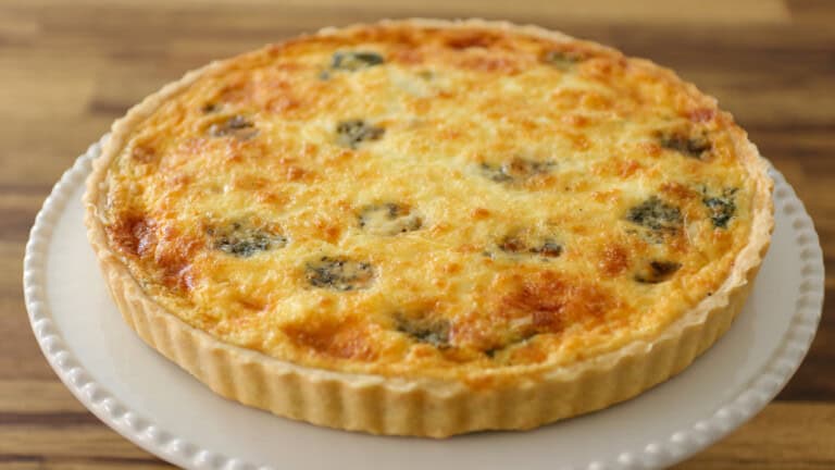 A golden-brown quiche with a flaky crust, topped with melted cheese and visible herbs, sits on a white plate atop a wooden surface.