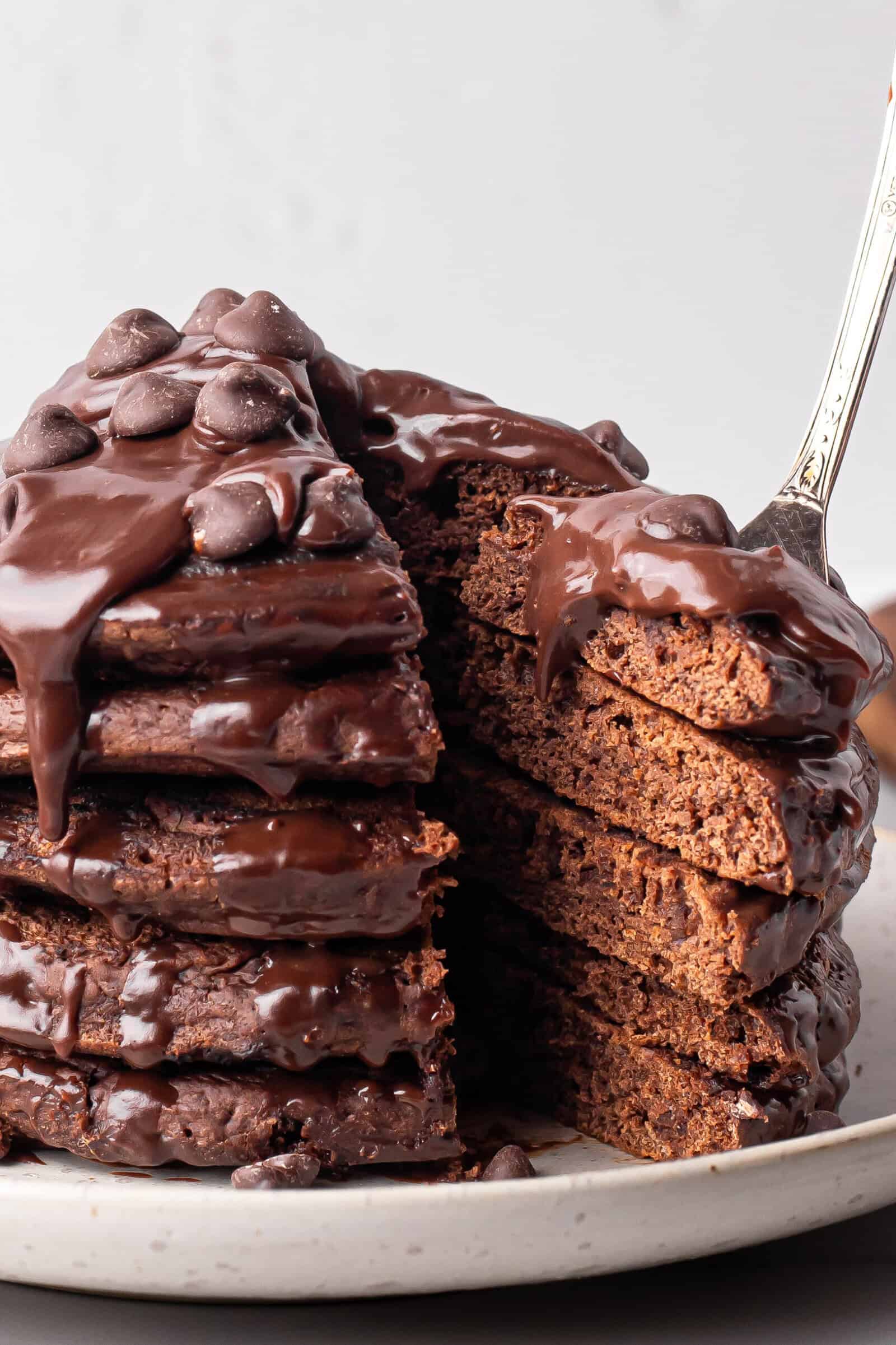 A stack of thick chocolate pancakes topped with melted chocolate sauce and chocolate chips, with a slice being lifted from the stack on a fork. The pancakes sit on a white plate against a light background.