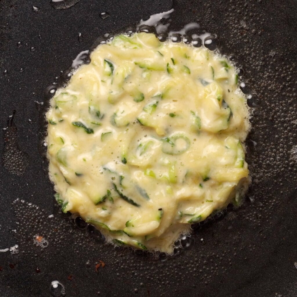 A zucchini fritter batter is cooking in a frying pan, with visible shredded zucchini and bubbles forming around the edges as it cooks in oil.
