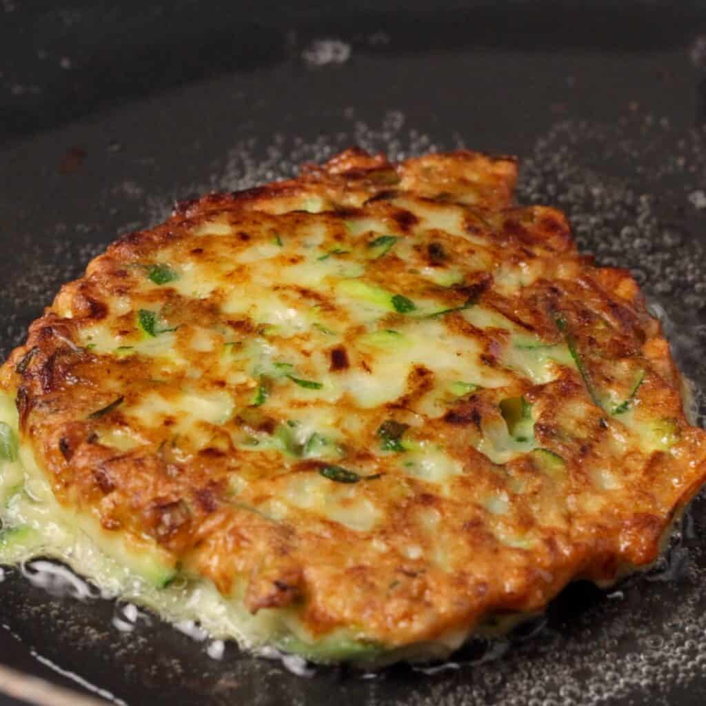 A golden-brown zucchini fritter with visible green herbs cooking in a black skillet, surrounded by sizzling oil.