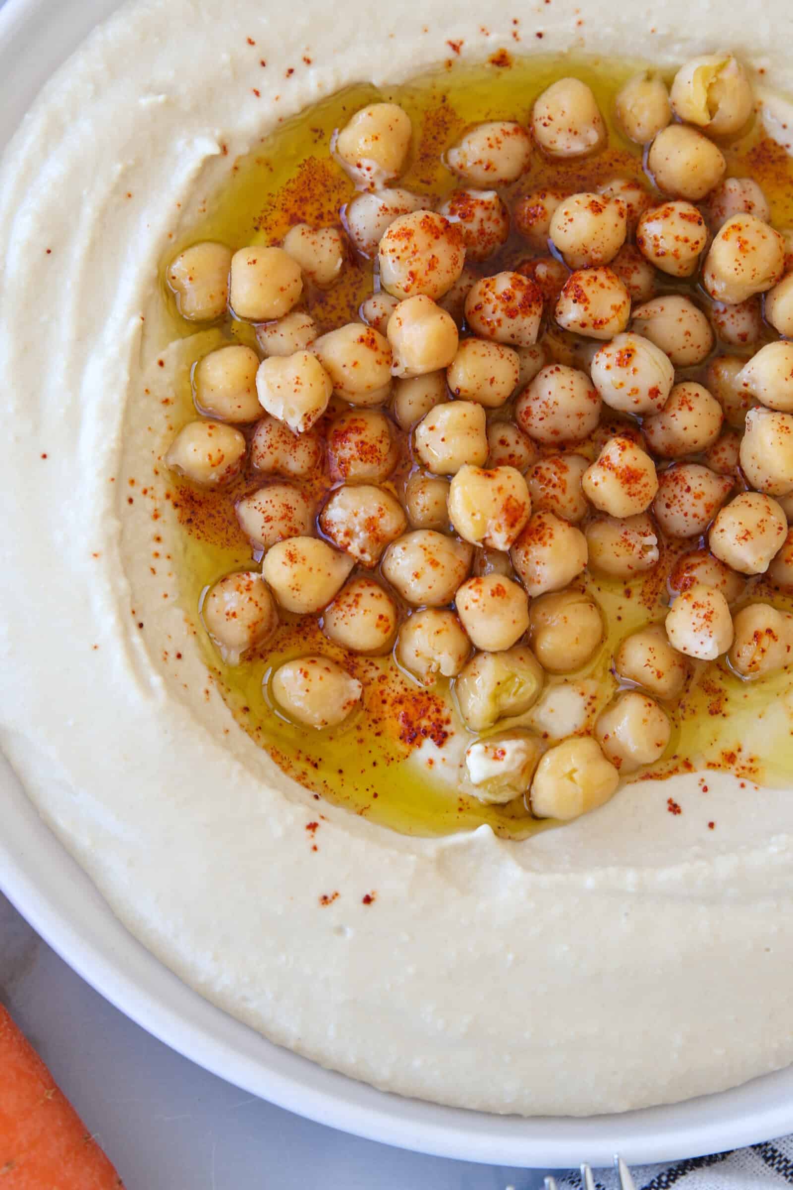 A dish of creamy hummus topped with whole chickpeas, a drizzle of olive oil, and a sprinkle of paprika, served in a white bowl.