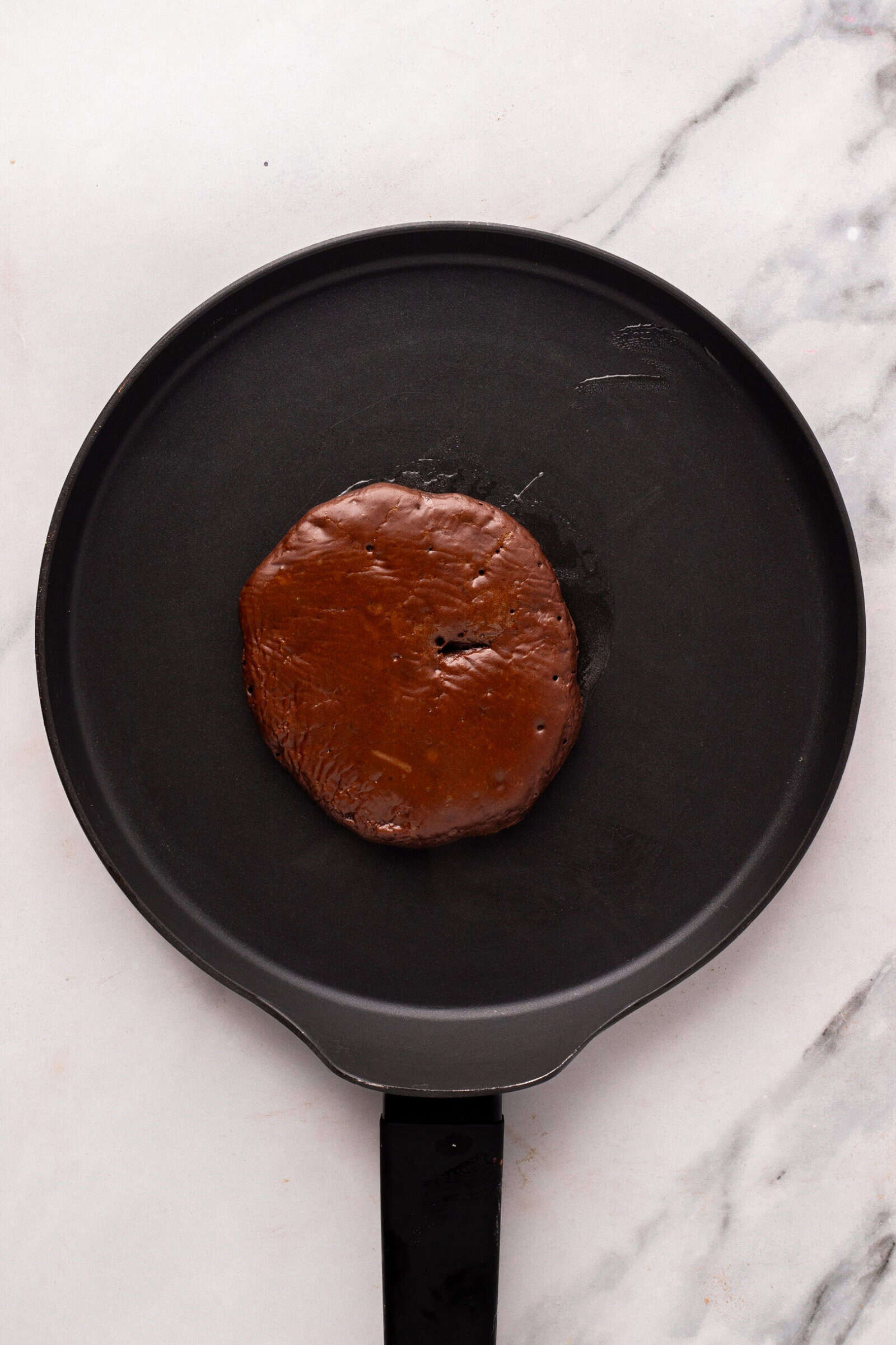 A round, dark chocolate pancake sits in the center of a black nonstick pan on a white marble surface.