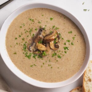 A bowl of creamy mushroom soup garnished with sautéed mushrooms and chopped parsley, served with slices of bread on the side.
