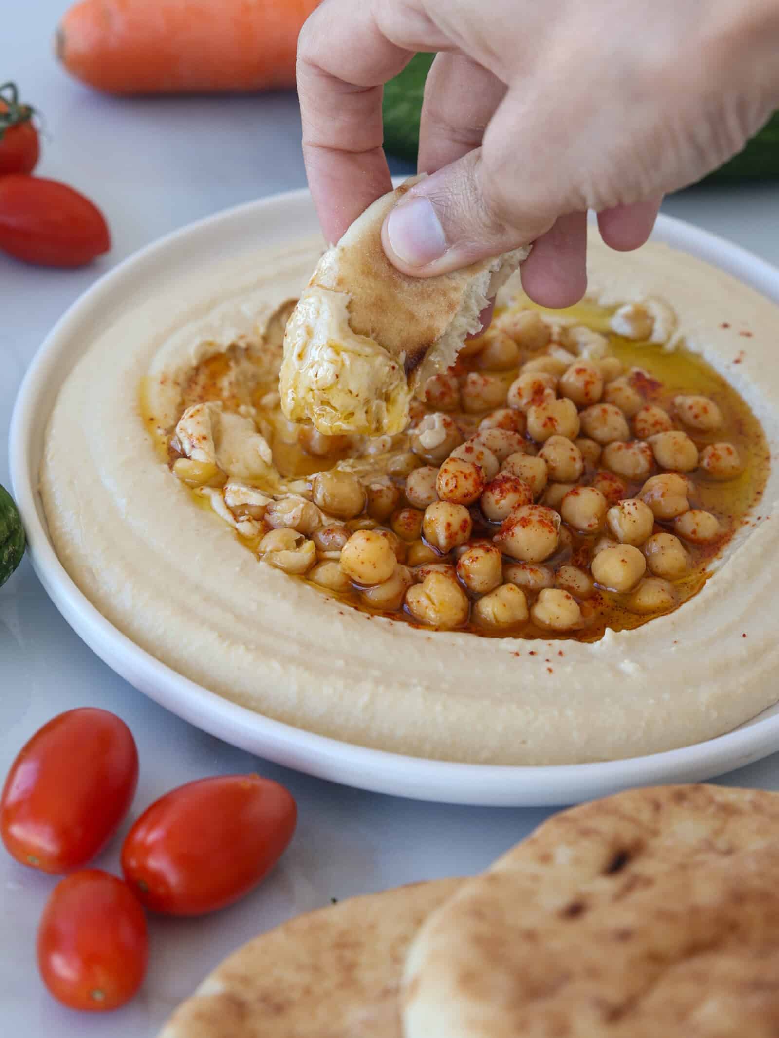 A hand dips a piece of pita bread into a bowl of creamy hummus topped with whole chickpeas, olive oil, and spices. Fresh vegetables and pita bread are visible around the plate.