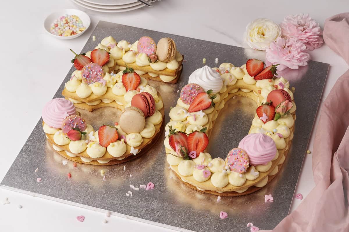 Large cookies shaped as the number 50 are decorated with cream, strawberries, macarons, meringues, and colorful sprinkles. The display is set on a silver board with flowers, plates, and a pink cloth nearby.