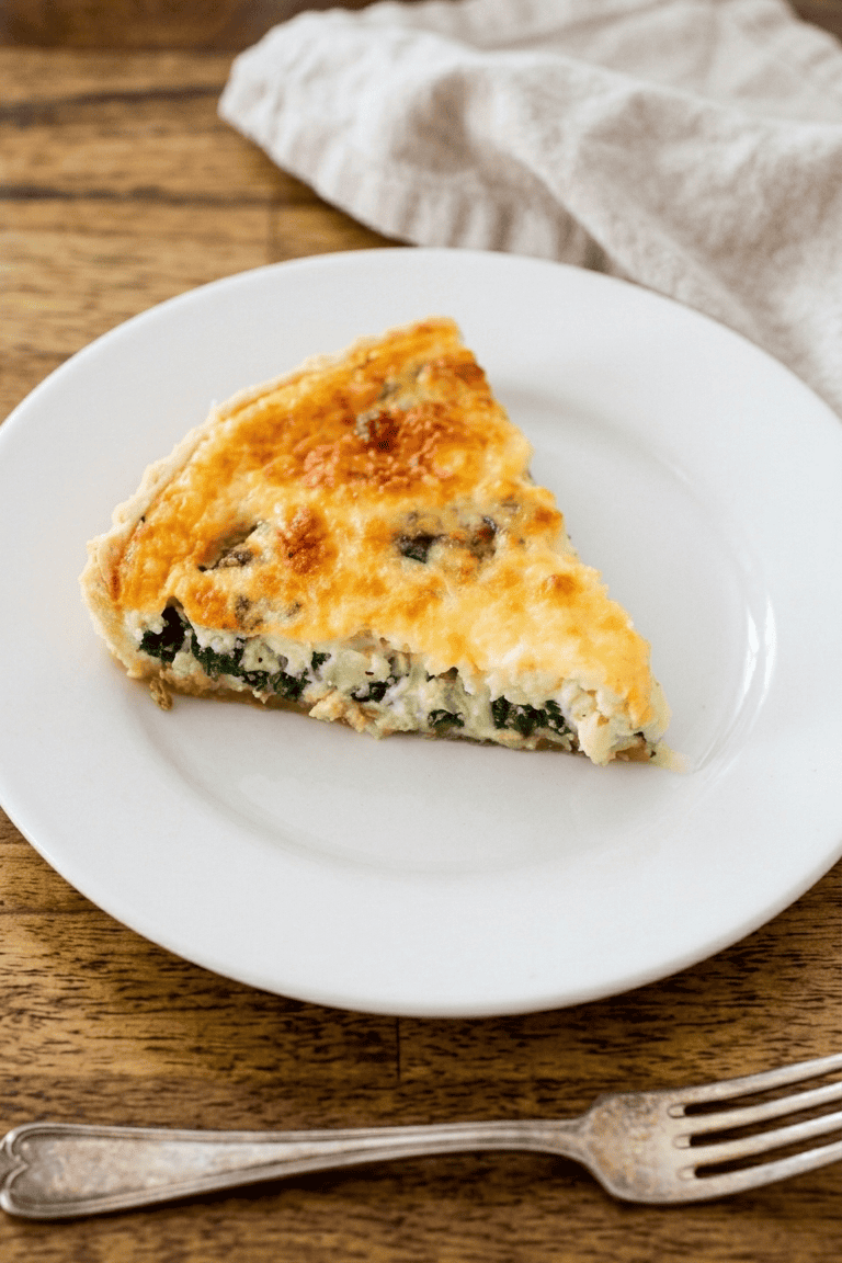 A slice of spinach and cheese quiche sits on a white plate, with a beige napkin and a fork beside it on a wooden table.