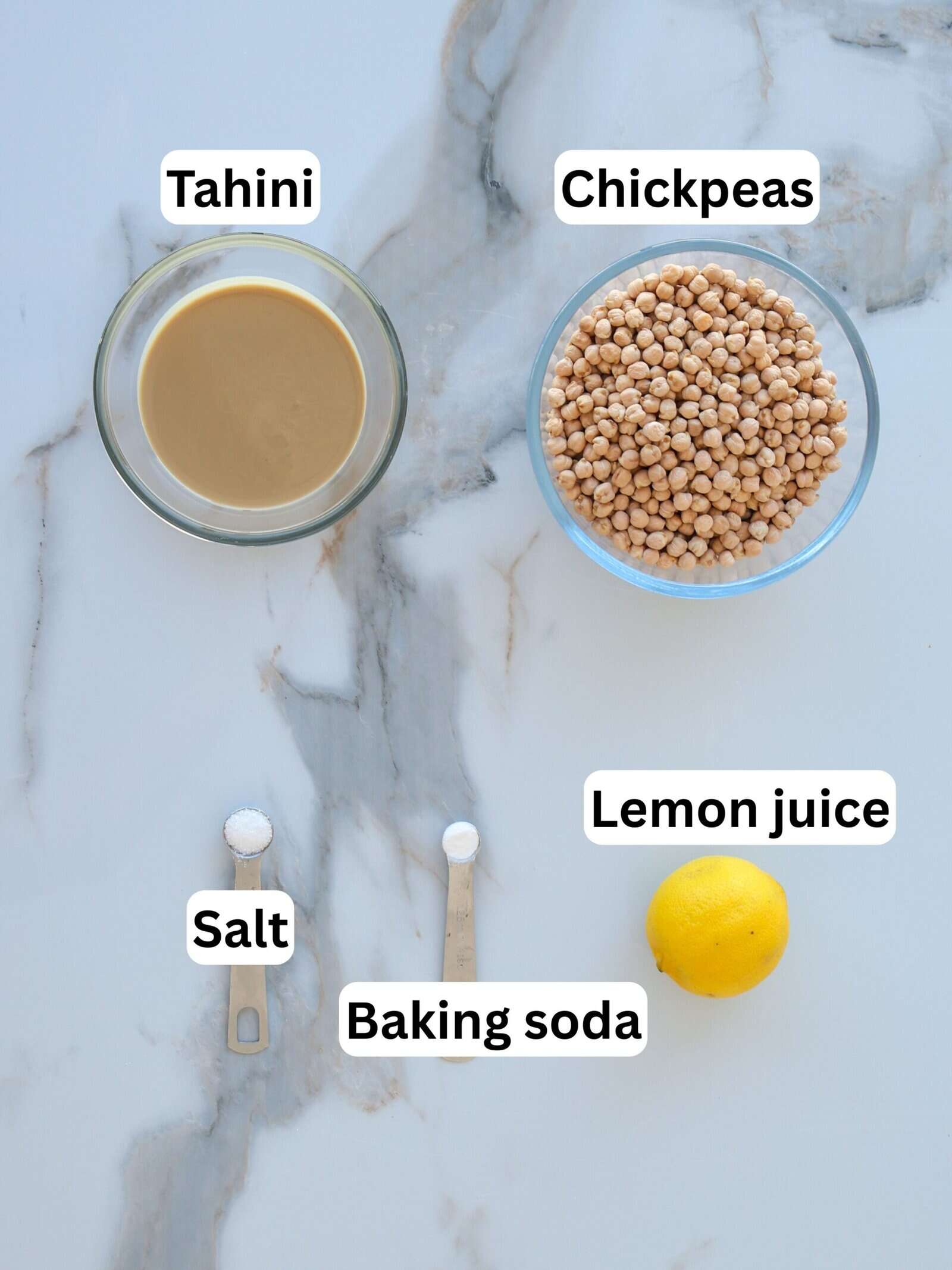 A bowl of tahini, a bowl of chickpeas, a whole lemon, and two measuring spoons containing salt and baking soda are arranged on a white marble surface. Each ingredient is labeled.