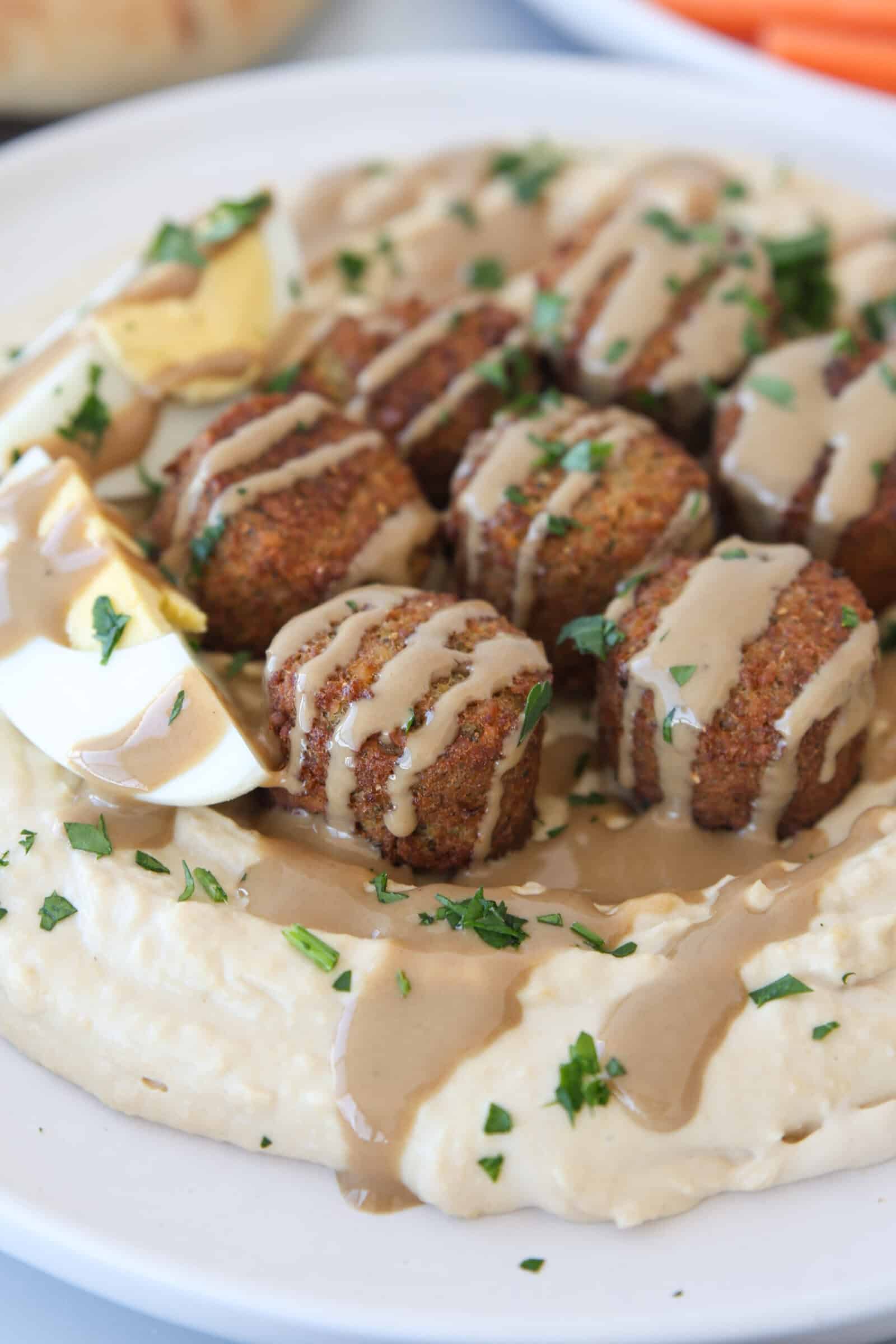 A plate of creamy hummus topped with crispy falafel balls, drizzled with tahini sauce, garnished with chopped parsley, and served with sliced hard-boiled eggs.