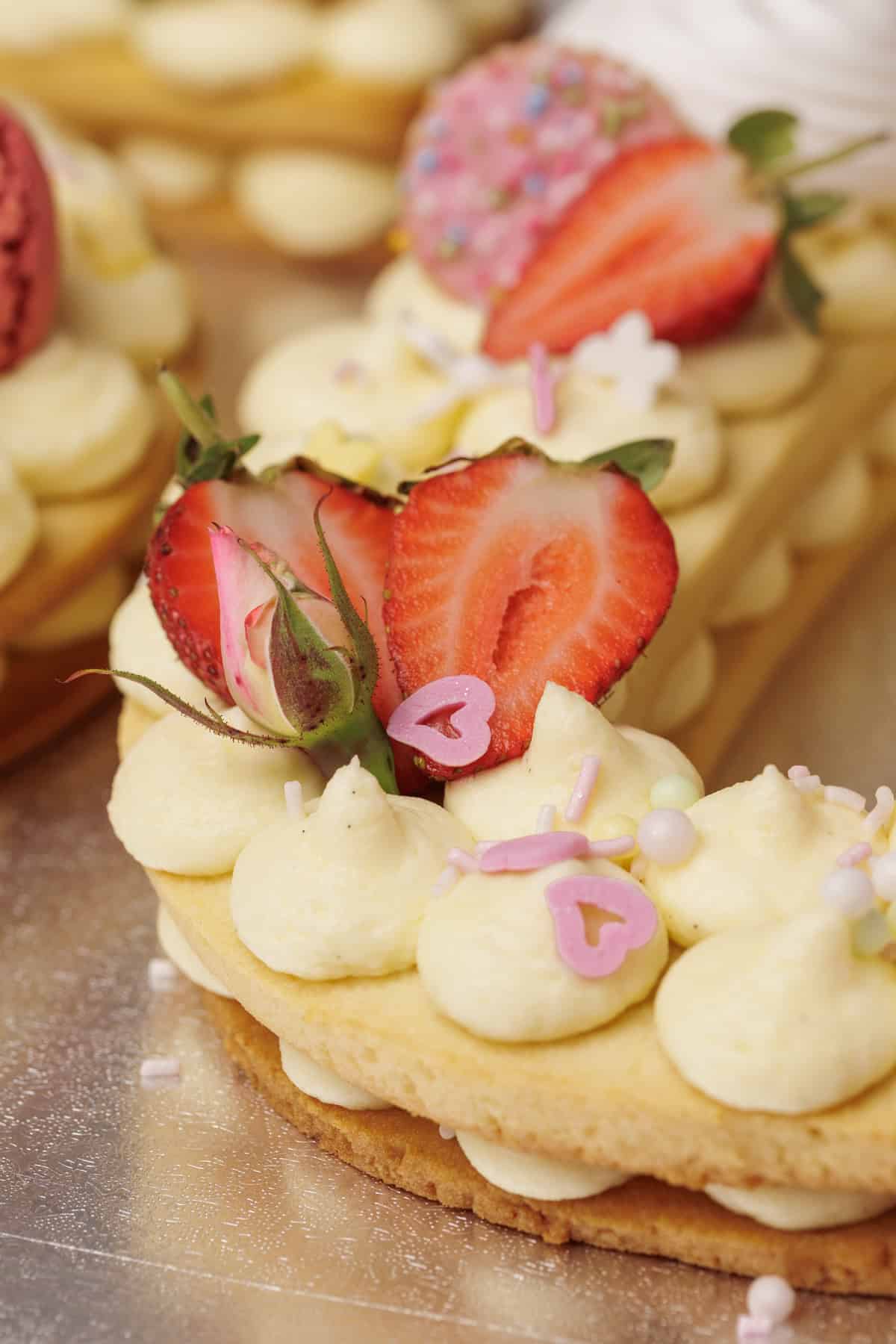 A close-up of a decorative number cookie cake topped with piped cream, sliced strawberries, a rosebud, heart-shaped sprinkles, and colorful decorations on a light-colored base.