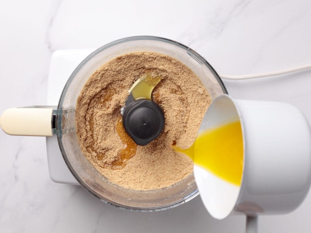 A food processor with crushed graham crackers inside is shown from above, while melted butter is being poured in from a white jug on a marble countertop.
