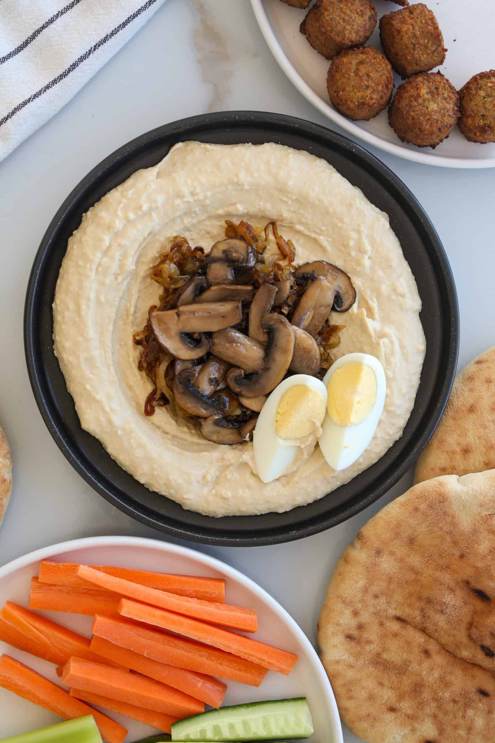 A plate of hummus topped with sautéed mushrooms, caramelized onions, and sliced boiled egg. Surrounding it are falafel, pita bread, cucumber and carrot sticks on a white surface.