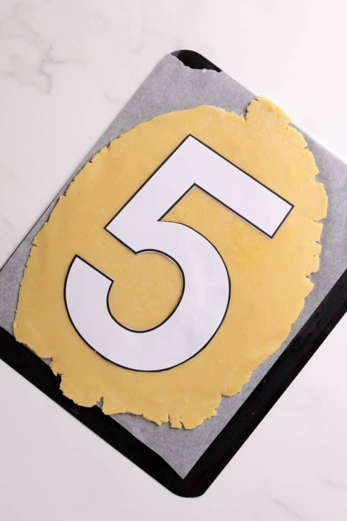 A large number 5 template lies on top of rolled-out cookie dough placed on a parchment-lined baking sheet.