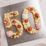 A large "50" number cake decorated with piped cream, strawberries, pink and beige macarons, and colorful sprinkles sits on a silver board, celebrating a milestone occasion.