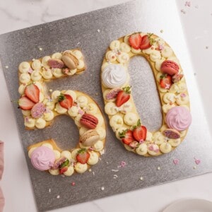 A large "50" number cake decorated with piped cream, strawberries, pink and beige macarons, and colorful sprinkles sits on a silver board, celebrating a milestone occasion.