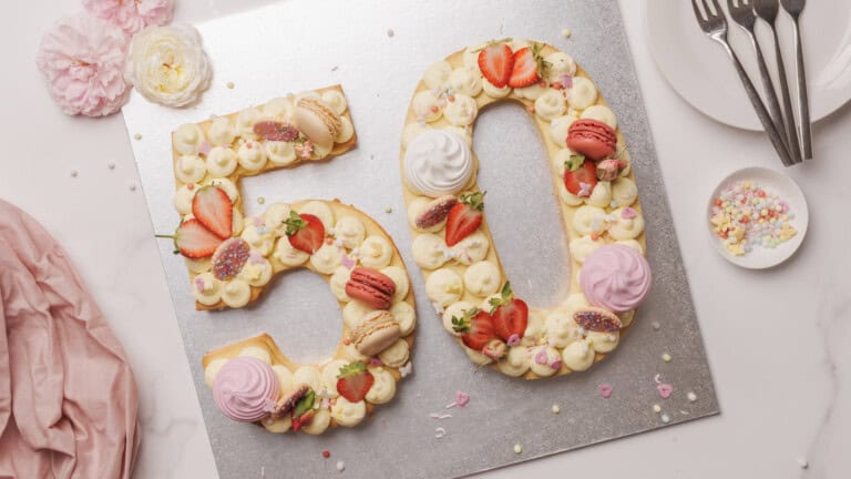 A cake shaped as the number 50 is decorated with strawberries, macarons, flowers, and piped frosting. The cake sits on a silver board near plates, forks, flowers, and a bowl of colorful sprinkles.