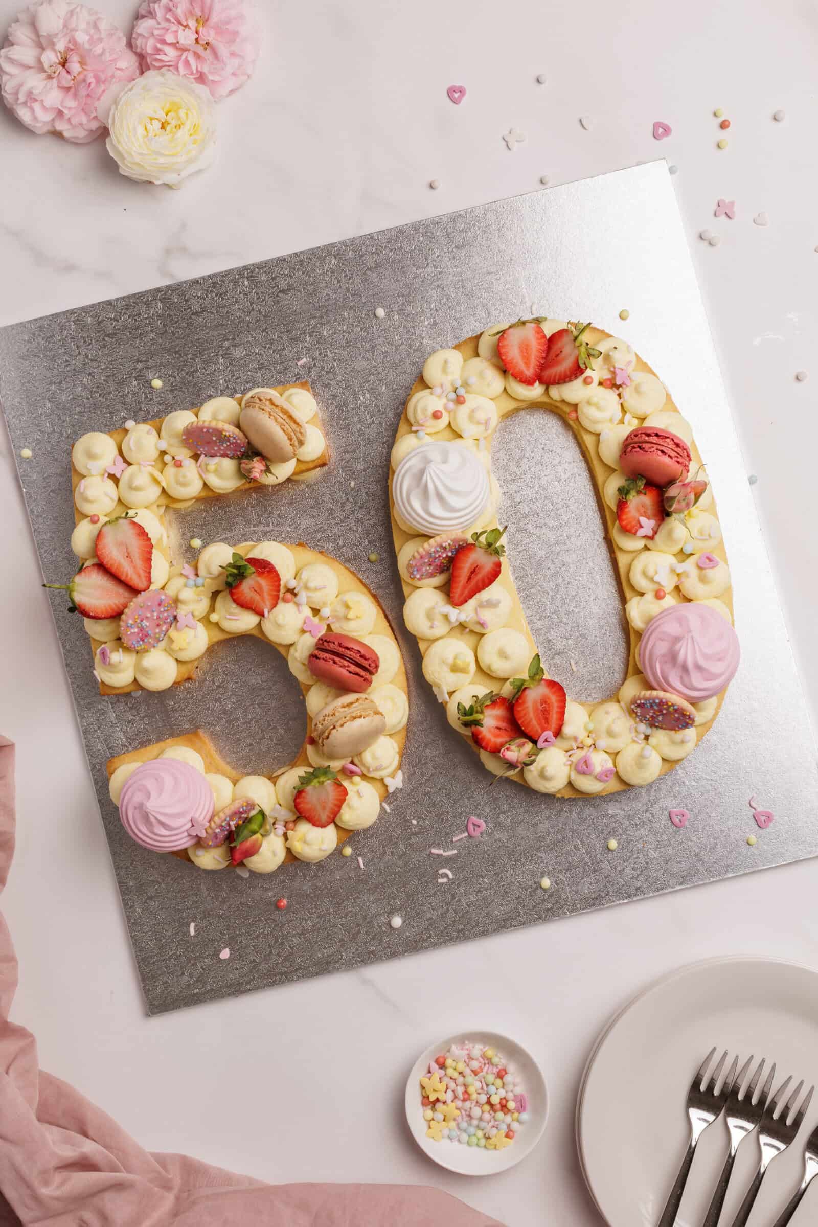 A cake shaped (number cake) as the number 50, decorated with strawberries, macarons, meringues, and cream, is displayed on a silver board. Plates, forks, sprinkles, and flowers are arranged nearby on a marble surface.