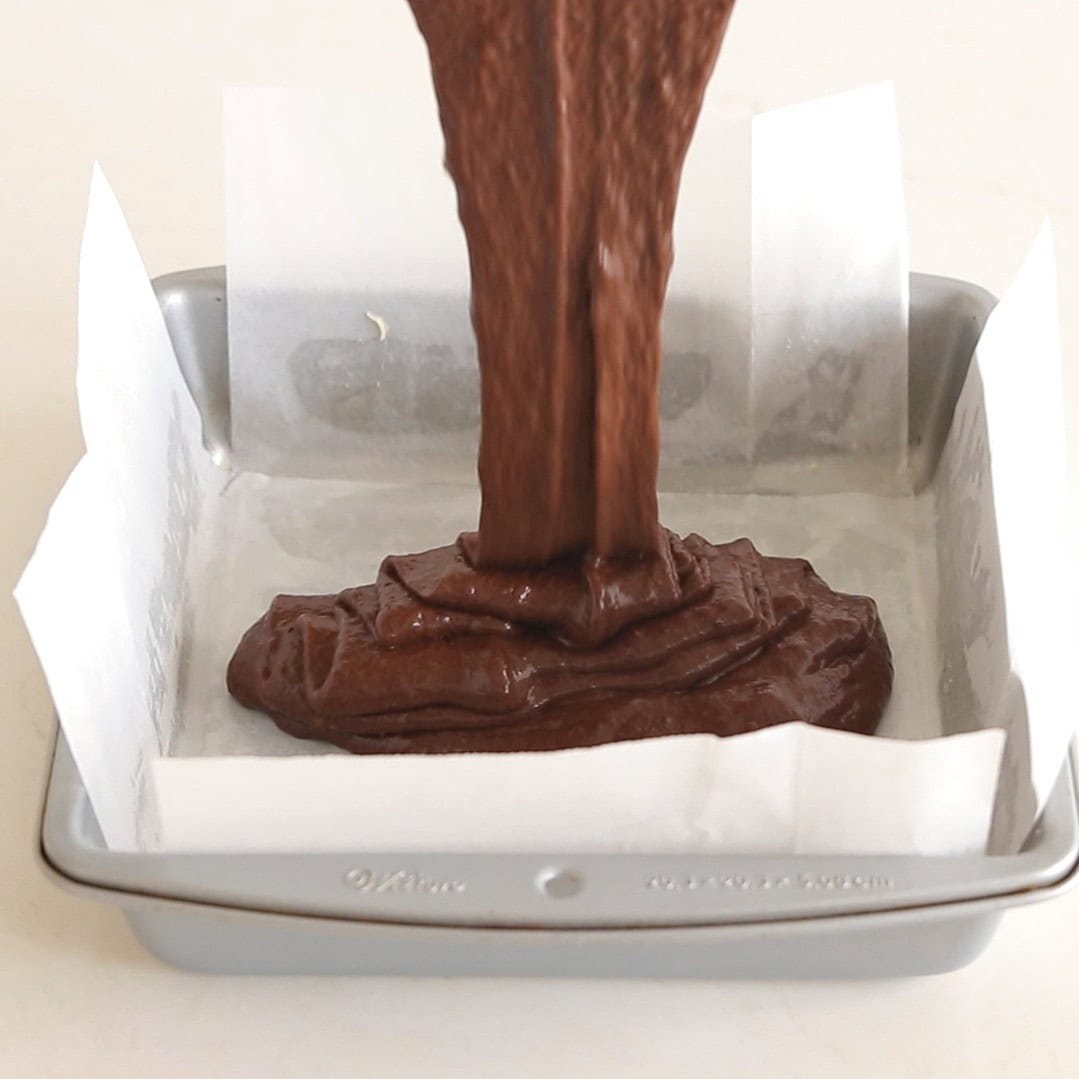 Thick chocolate batter is being poured into a square baking pan lined with parchment paper, ready for baking.
