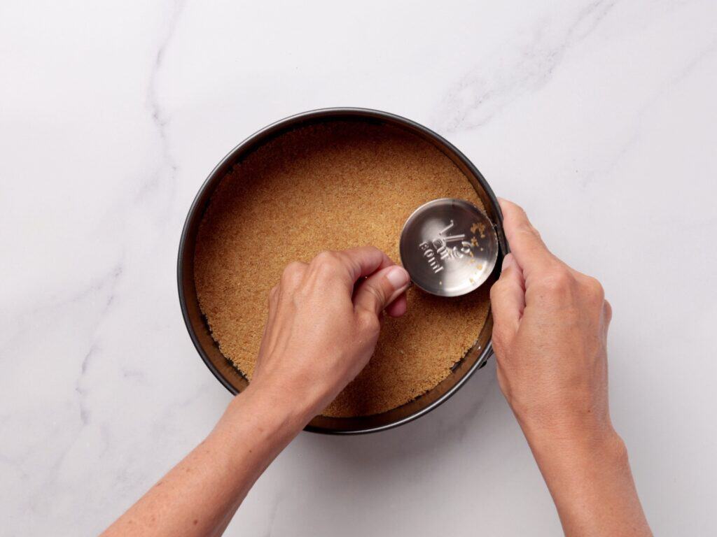 A person presses down a crumb mixture in a round baking pan using a metal measuring cup on a white marble surface.