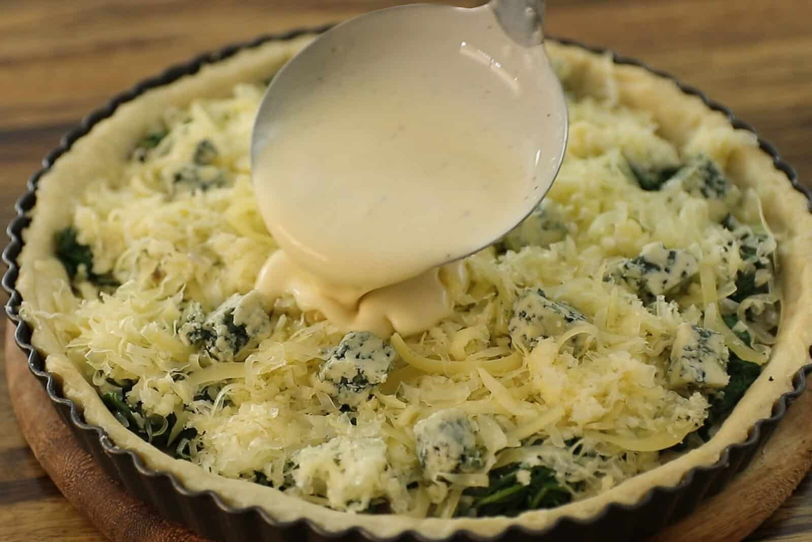 A close-up of a quiche being prepared; a spoon pours creamy egg mixture over a crust filled with shredded cheese, spinach, and blue cheese, set on a wooden surface.