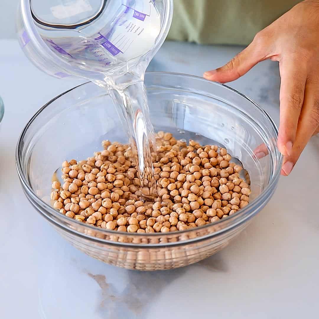 A person pours water from a measuring cup into a glass bowl filled with dry chickpeas on a white surface.