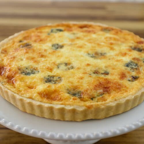 A freshly baked spinach and cheese quiche with a golden, cheesy top and a flaky crust sits on a white cake stand against a wooden background.