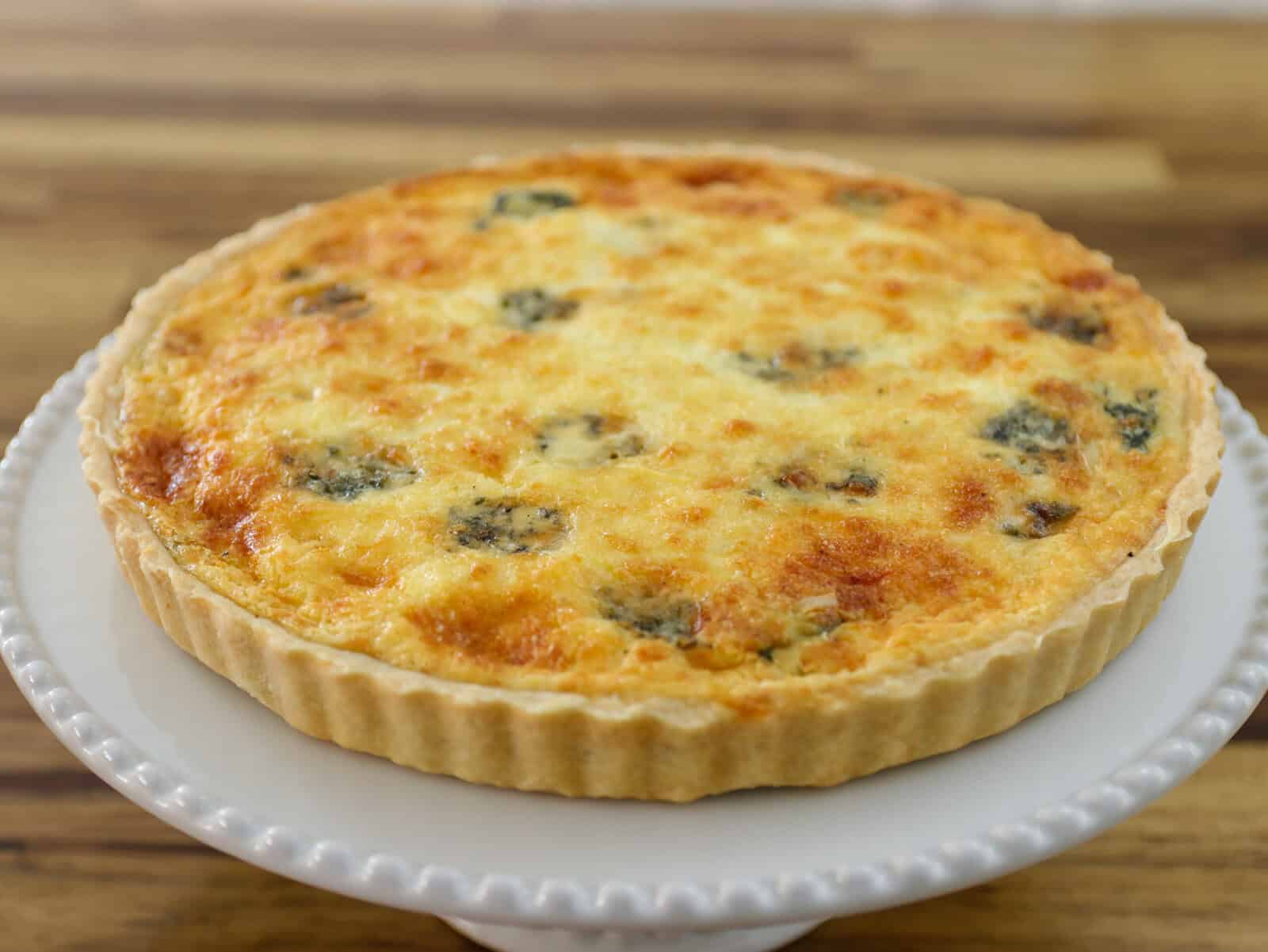 A freshly baked spinach and cheese quiche with a golden, cheesy top and a flaky crust sits on a white cake stand against a wooden background.