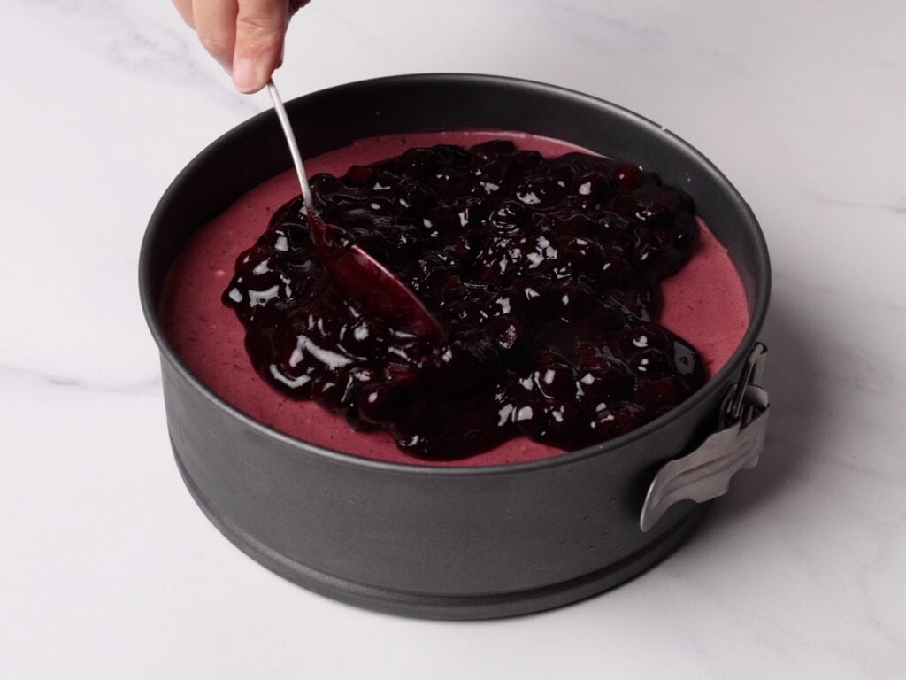 A hand uses a spoon to spread a thick layer of blueberry topping over a purple cheesecake mixture in a round springform pan on a white surface.