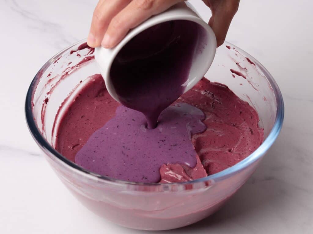 A hand pours thick, purple batter from a white cup into a glass mixing bowl already containing a similar purple mixture, on a white surface.
