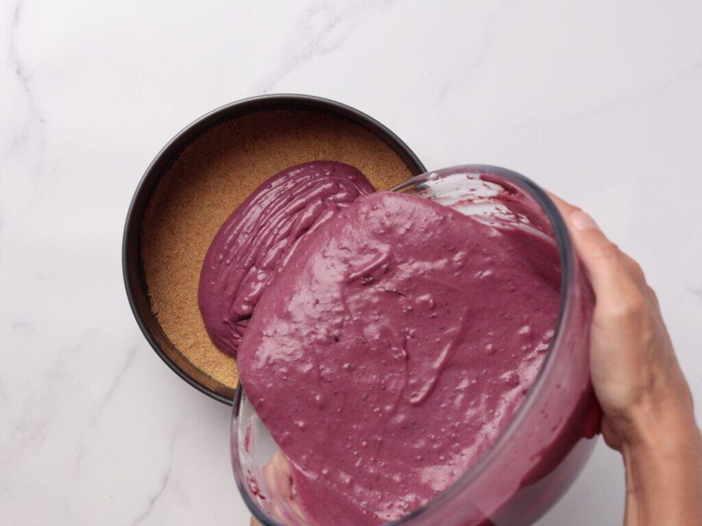 A hand pours thick purple cake batter from a glass bowl into a round pan lined with a brown crust, on a white marble surface.