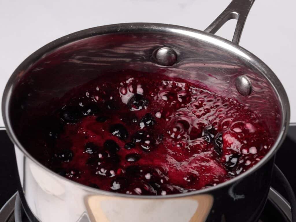 A stainless steel saucepan on a stove containing bubbling, dark purple berry sauce with visible whole berries, likely being cooked or reduced.