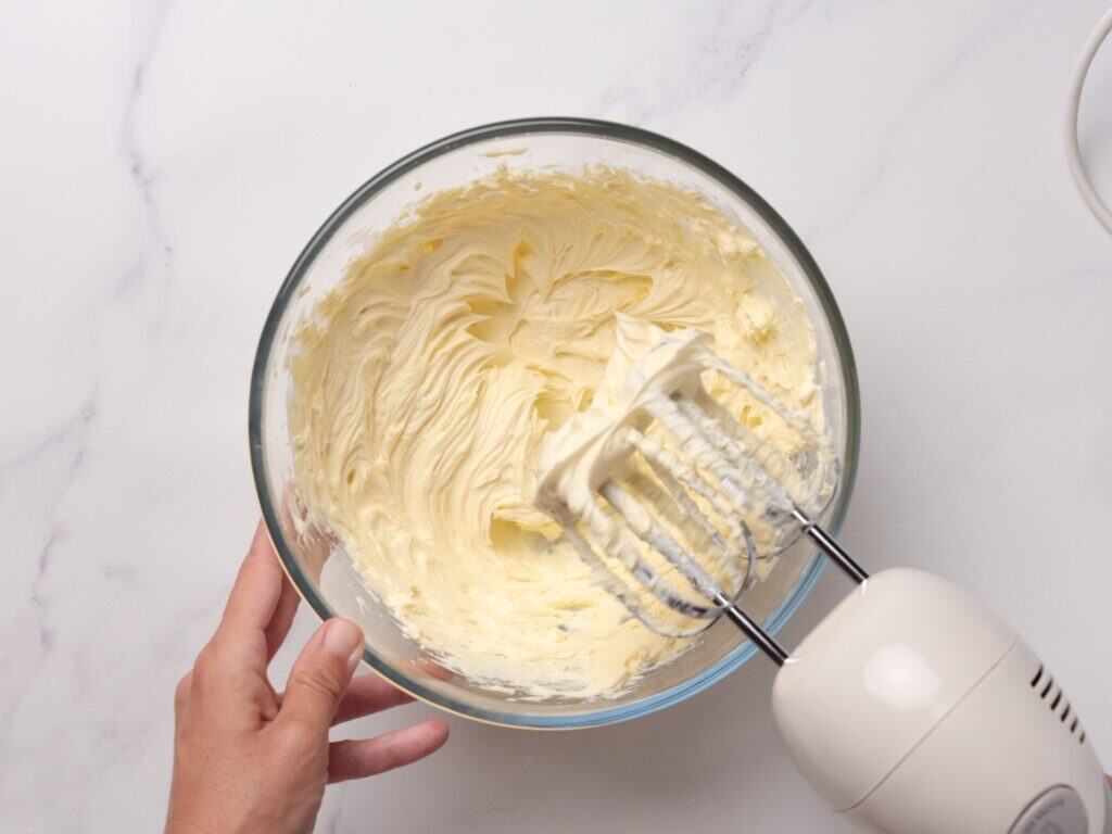 A hand holds a glass bowl of whipped buttercream while an electric hand mixer with beaters is mixing the creamy mixture on a white countertop.