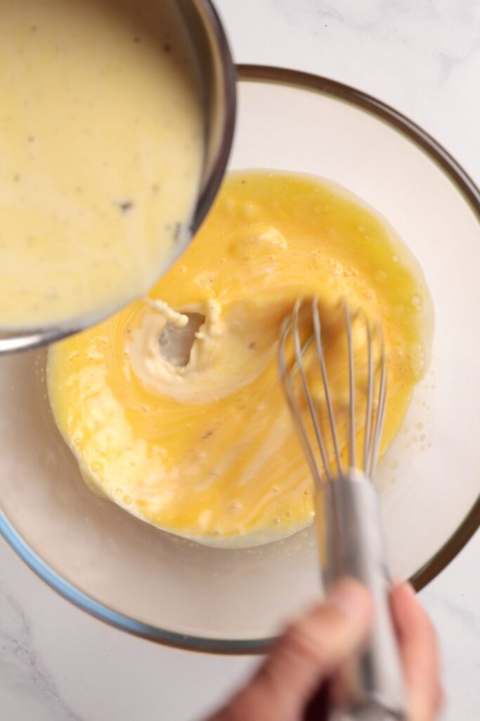 A hand whisks eggs in a glass bowl while creamy sauce is being poured in from a saucepan, blending the ingredients together on a white countertop.