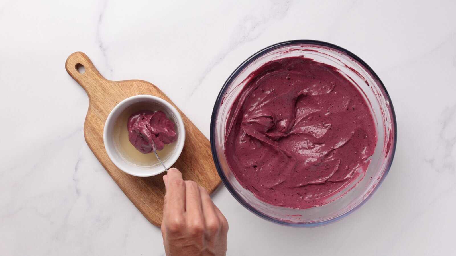 A hand scoops thick, purple batter from a glass bowl into a small white bowl on a wooden board, all set on a white marble surface.