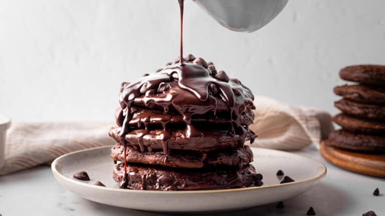 A stack of chocolate pancakes topped with chocolate chips is being drizzled with thick chocolate sauce, sitting on a white plate with a neutral background.