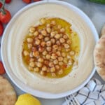 A bowl of creamy hummus topped with whole chickpeas, olive oil, and paprika, surrounded by pita bread, tomatoes, a lemon, and a striped cloth napkin.