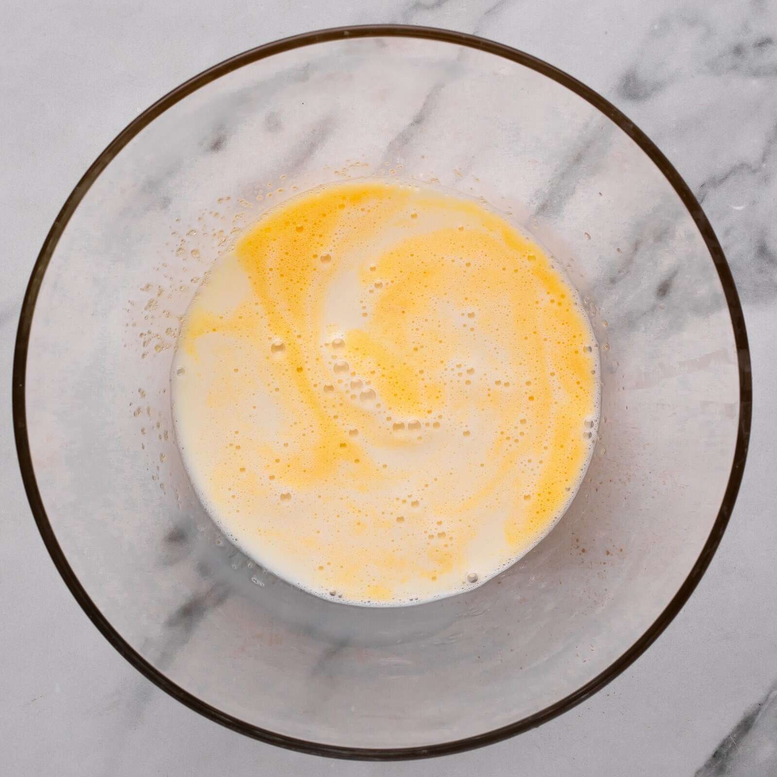 A glass bowl on a marble surface contains a mixture of beaten eggs and milk, showing a pale yellow and white marbled appearance.