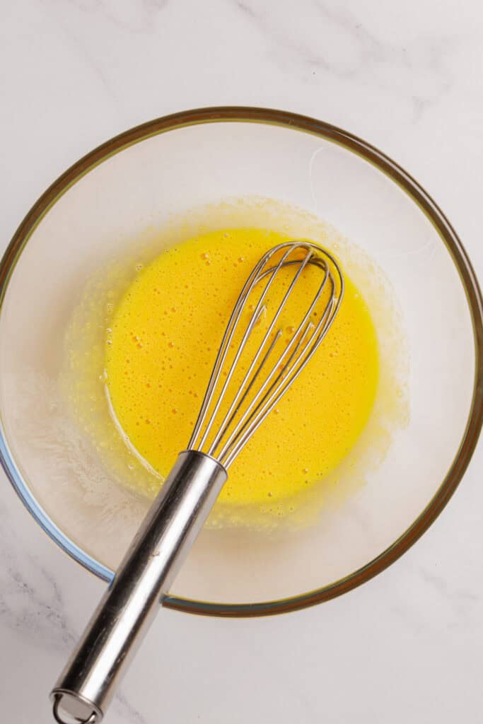 A metal whisk rests in a glass bowl filled with a beaten yellow egg mixture, placed on a white marble surface.