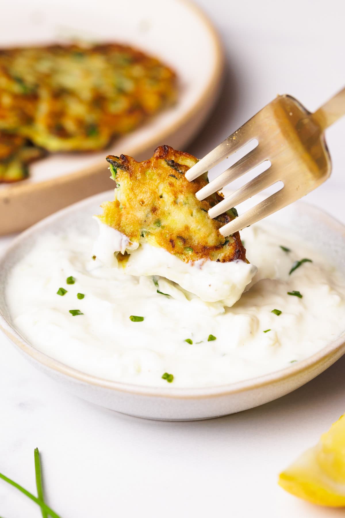 A fork holds a zucchini fritter dipped in creamy white sauce, garnished with chopped herbs. In the background, more fritters rest on a plate. The scene is bright and appetizing.