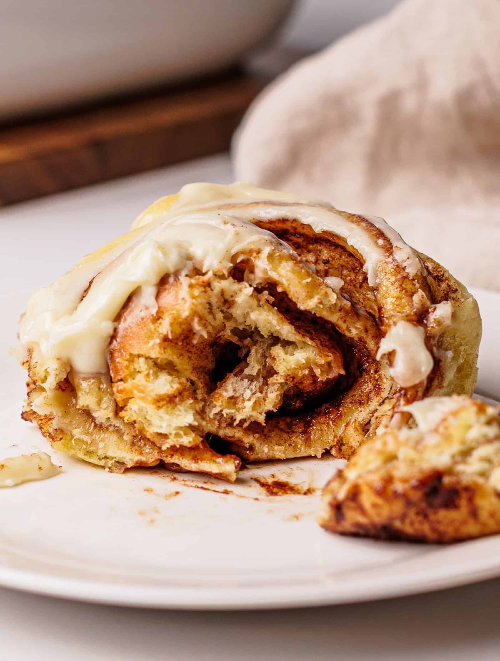 A close-up of a partially eaten cinnamon roll with cream cheese icing on a white plate, showing its soft, swirled layers. A small piece is separated in the foreground.